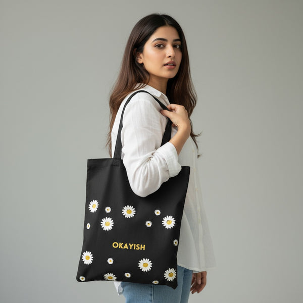 Woman holding a black tote bag with white floral patterns and 'OKAYISH' text on a gray background